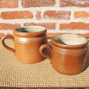 May include: Two cylindrical mugs with a warm brown exterior glaze and a smooth, light-colored interior. Each mug features a sturdy, curved handle attached to the side and a distinct raised rim around the top edge, showcasing a simple, functional design.