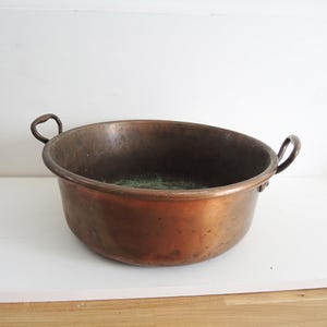 以下が含まれることがあります： 2つの湾曲したハンドルが付いた、大きなアンティークの銅製洗面器。洗面器は幅が広く浅いボウルで、赤褐色の豊かな古色を帯びています。内部には、緑色の酸化による経年変化と使用の兆候が見られます。