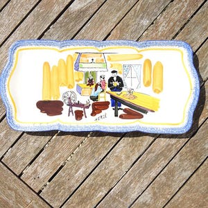 May include: Rectangular ceramic serving platter with a scalloped edge, featuring a hand-painted scene of a bakery. The border is blue and yellow. The scene includes figures, a table, and baked goods. The word "HERCE" is written on the platter.