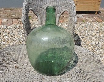 Puede incluir: Una gran botella de vidrio verde con cuerpo redondo y cuello estrecho. La botella está sentada en una mesa de mimbre blanca.