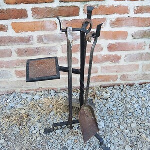 May include: A black metal fireplace tool set with a shovel, poker, and brush. The tools are mounted on a stand with a square platform. The set is in front of a brick wall.