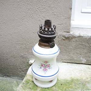 May include: A vintage oil lamp with a white ceramic base, adorned with pink rose floral patterns and blue trim. The lamp features a brass burner and a decorative metal crown. A classic decorative piece.