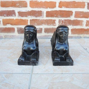 Peut inclure: Une paire de figurines sphinx en fonte noire, chacune présentant une tête humaine détaillée avec des cheveux stylisés et un corps félin. Ces statues en métal massif et lourd sont conçues avec une base rectangulaire et une finition sombre.
