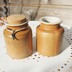 May include: Two ceramic jars with a rustic appearance. One jar has a cork stopper secured with twine and black details. The other jar is open, showing a white interior. Both jars are a warm, earthy brown, suggesting a handmade quality.