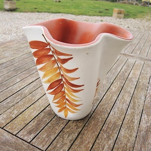 May include: A large, off-white ceramic vase with a wavy rim and a reddish-orange interior. The exterior features a painted design of brown and yellow-orange fern leaves. The vase is set on a wooden surface.