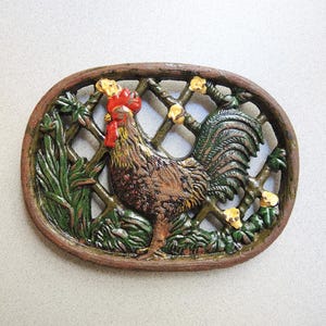 May include: Oval-shaped, cast iron rooster wall decor. The rooster is brown, red, and green, set against a lattice and floral background. The border is a dark brown. A decorative piece for a kitchen or farmhouse-style home.