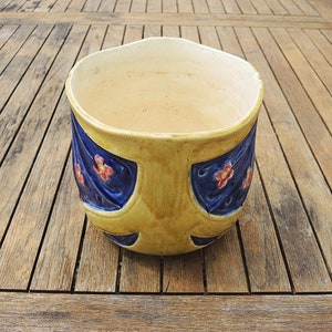 May include: A yellow ceramic planter with blue and pink floral designs. The planter has a rounded shape and a wide opening.