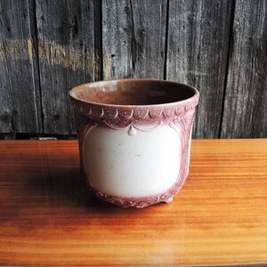 May include: A vintage, ceramic planter with a white center and a decorative, scalloped rim in a muted burgundy color. The planter has a rounded shape with a slightly textured surface and sits on small feet. The background is a weathered wooden wall.