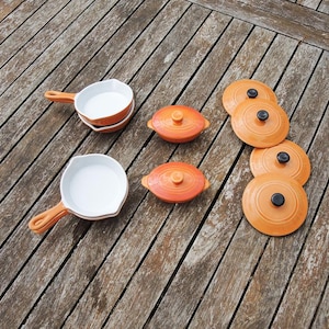Puede incluir: Juego de utensilios de cocina de cerámica naranja con interiores blancos. El juego incluye pequeñas cacerolas con asas, platos ovalados cubiertos y tapas a juego con pomos negros. Los artículos están dispuestos sobre una superficie de madera envejecida.