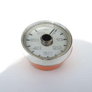 Può includere: Un misuratore circolare Jaeger con quadrante bianco, marcature numeriche nere da 10 a 60 e un'apertura centrale nera. Lo strumento presenta una lunetta in metallo argentato lucido e un alloggiamento di base cilindrico color arancione.