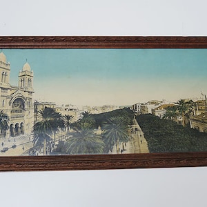 May include: A framed vintage postcard depicting a cityscape with a large church building in the foreground. The church has two tall towers and a white facade. The city street is lined with palm trees and buildings. The postcard is in a brown wooden frame.