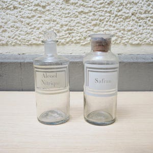 Puede incluir: Dos botellas de vidrio transparente con tapones de corcho y etiquetas blancas. La botella de la izquierda tiene el texto "Alcool Nitrique" en la etiqueta. La botella de la derecha tiene el texto "Safran" en la etiqueta.