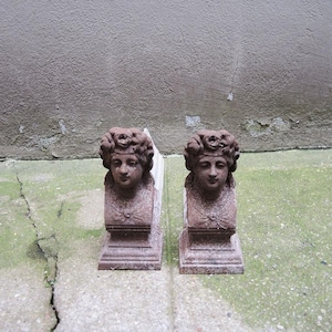 Puede incluir: Dos sujetalibros antiguos, cada uno con un busto esculpido de mujer con cabello adornado y una corona floral. Los sujetalibros están hechos de un material marrón rojizo desgastado y descansan sobre una base rectangular. El fondo es una pared gris texturizada.