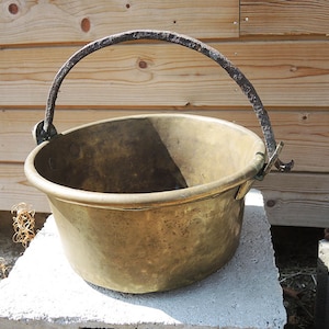 May include: A large, antique brass pot with a rusty iron handle. The pot is sitting on a concrete block.
