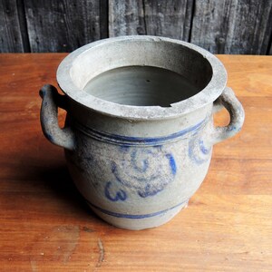 May include: A gray stoneware crock with blue painted designs and two handles. The crock is sitting on a wooden surface.