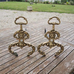 Puede incluir: Un par de morillos de chimenea antiguos de latón. Cada morillo presenta una cabeza de león decorativa, adornos ornamentados y un asa. Los morillos se exhiben sobre una mesa de madera al aire libre.