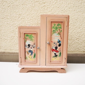 May include: A small, pink wooden cabinet featuring hand-painted images of Mickey and Minnie Mouse. The cabinet has two doors, each with a framed illustration of the characters, trees, and flowers. The cabinet is a vintage piece.