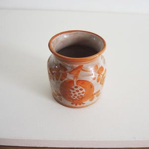 Puede incluir: Un pequeño jarrón de cerámica hecho a mano con una abertura ancha y un cuerpo redondeado. El jarrón presenta una base de color crema con diseños florales y frutales de color naranja. El borde y el interior son de color naranja oscuro.
