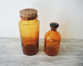 May include: Two amber glass bottles with cork and black glass stoppers. The larger bottle is taller and has a cork stopper. The smaller bottle is shorter and has a black glass stopper.