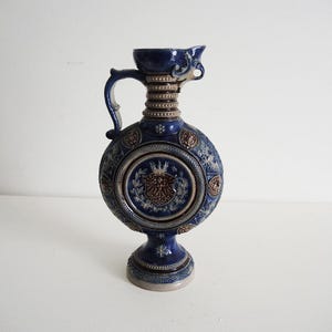 May include: A blue ceramic pitcher with a handle and spout. The pitcher has a round body with a detailed emblem and floral designs. The base and neck feature additional decorative elements.
