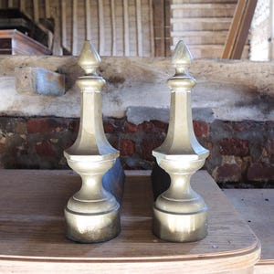 Op de afbeelding: Twee antieke messing en bronzen haardijzers met een decoratief, geometrisch ontwerp. Elk haardijzer heeft een conische top, een facetvormig lichaam en een ronde basis. Ze worden gepresenteerd op een houten oppervlak.