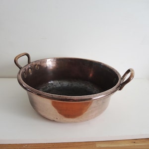 Op de afbeelding: Een vintage koperen kom met twee gebogen handvatten. De kom heeft een breed, ondiep ontwerp met een donker interieur en een gepolijste buitenkant. Het koper heeft een warme, roodbruine tint en de handvatten zijn van een lichter metaal.