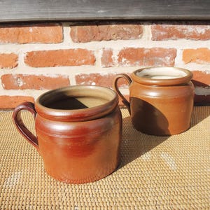 May include: Two brown ceramic mugs with a smooth, glazed finish and a rounded, sturdy handle on each side. The mugs feature a wide, cylindrical body with a thick, rolled rim at the top and a light-colored interior, showcasing a simple design.
