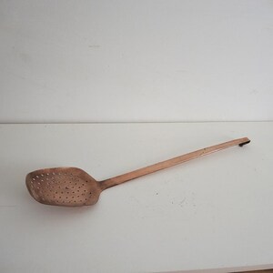 May include: A copper-colored slotted spoon with a long handle. The spoon's head is rectangular with numerous small holes. The handle is straight and tapers slightly towards the end. The spoon is made of metal and appears to be vintage.