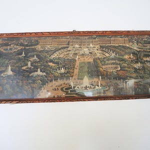 May include: An antique framed print depicting a detailed aerial view of a European city park with fountains, buildings, and trees. The print is encased in a dark brown, carved wooden frame, adding a vintage aesthetic.