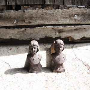 Puede incluir: Dos bustos de piedra envejecida de una persona joven, posiblemente Juana de Arco, con pelo corto y una coraza detallada. Los bustos son de color gris desgastado, con textura y detalles visibles. El fondo es una superficie de madera envejecida.