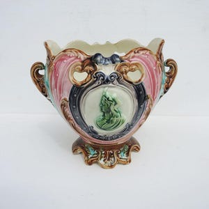 May include: A ceramic jardiniere featuring a central oval medallion with a green relief profile of a person. The vessel is decorated with pink, brown, and teal glazes, ornate scrollwork, two side handles, and a scalloped rim and base edge.