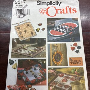 May include: A Simplicity 9517 sewing pattern envelope for various fabric crafts. The front displays images of a quilted wall hanging, a checkerboard runner, a backgammon board, a sunflower ornament, a tic-tac-toe game, and apple-themed decor.