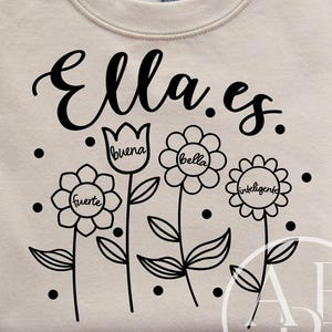 May include: A cream-coloured sweatshirt featuring the words "Ella es" in black script. Below the text are four flower illustrations, each with a word inside: "fuerte", "buena", "bella", and "inteligente". The sweatshirt is folded.