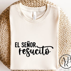 Op de afbeelding: Crèmekleurig T-shirt met de Spaanse tekst "EL SEÑOR resucitó" in zwart script. Het shirt wordt gepresenteerd op een geweven, lichtbruin oppervlak. Het ontwerp bevat een cirkelvormig logo in de rechter benedenhoek.
