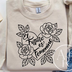 以下が含まれることがあります： ベージュのスウェットシャツに黒いグラフィック。グラフィックは「El Futuro es Femenino」という言葉が入ったハートで、バラと葉で囲まれています。スウェットシャツは白い表面に折りたたまれています。