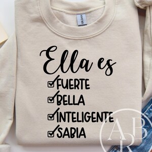 May include: A beige sweatshirt featuring the phrase "Ella es" in black script. Underneath, the words "FUERTE," "BELLA," "INTELIGENTE," and "SABIA" are listed with checkmarks. The sweatshirt is folded, with jeans and a coffee cup visible.