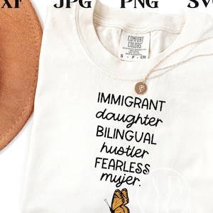 May include: Cream-colored t-shirt with the words "IMMIGRANT daughter BILINGUAL hustler FEARLESS mujer" in black script. A small butterfly graphic is at the bottom. A gold necklace is draped on the shirt.