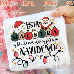 May include: White t-shirt with a Christmas-themed design. The design includes a Santa Claus illustration, Christmas ornaments, and text in Spanish that reads "Esta Maestra está llena de espíritu navideño."