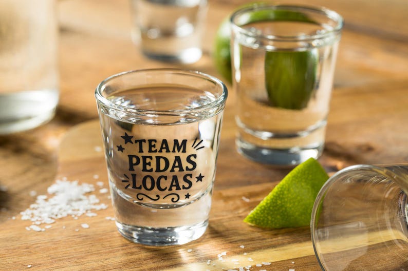K&ouml;nnte beinhalten: Shotgl&auml;ser gef&uuml;llt mit klarer Fl&uuml;ssigkeit, mit dem Text "TEAM PEDAS LOCAS" in Schwarz. Die Gl&auml;ser stehen auf einer Holzoberfl&auml;che, mit Limettenspalten und Salz in der N&auml;he. Die Shotgl&auml;ser sind klar und zylindrisch.