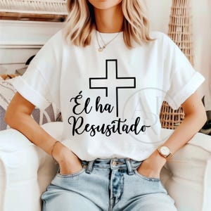 Puede incluir: Camiseta blanca con una cruz negra y la frase en español "Él ha Resucitado". La camiseta tiene mangas cortas y un corte holgado. El diseño es sencillo y elegante, adecuado para uso casual.