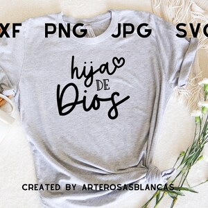 Puede incluir: Camiseta gris con el texto "hija de Dios" en letra negra cursiva con un peque&ntilde;o coraz&oacute;n sobre la palabra "hija". El texto est&aacute; centrado en la camiseta. La camiseta est&aacute; colocada sobre una superficie blanca con una cesta tejida y flores blancas.