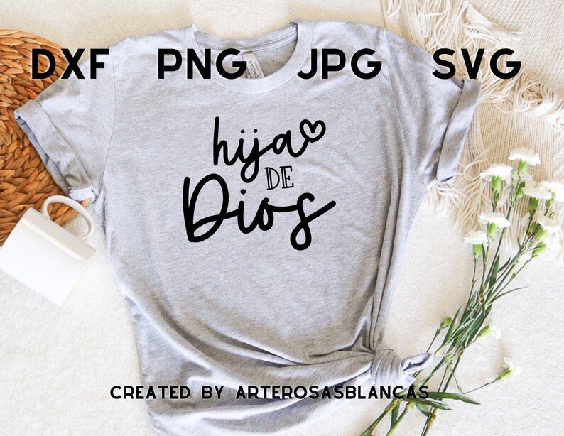 Puede incluir: Camiseta gris con el texto "hija de Dios" en letra negra cursiva con un peque&ntilde;o coraz&oacute;n sobre la palabra "hija". El texto est&aacute; centrado en la camiseta. La camiseta est&aacute; colocada sobre una superficie blanca con una cesta tejida y flores blancas.