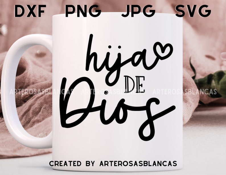 Puede incluir: Taza de cer&aacute;mica blanca con un dise&ntilde;o negro que dice "hija de Dios" con un peque&ntilde;o coraz&oacute;n al lado de la palabra "hija". La taza est&aacute; sobre una superficie de tela rosa.
