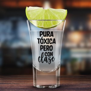 K&ouml;nnte beinhalten: Ein Shotglas gef&uuml;llt mit einer klaren Fl&uuml;ssigkeit, garniert mit einer Limettenscheibe. Auf dem Glas steht der Schriftzug "Pura T&oacute;xica Pero Con Clase". Das Shotglas steht auf einer Holzoberfl&auml;che.