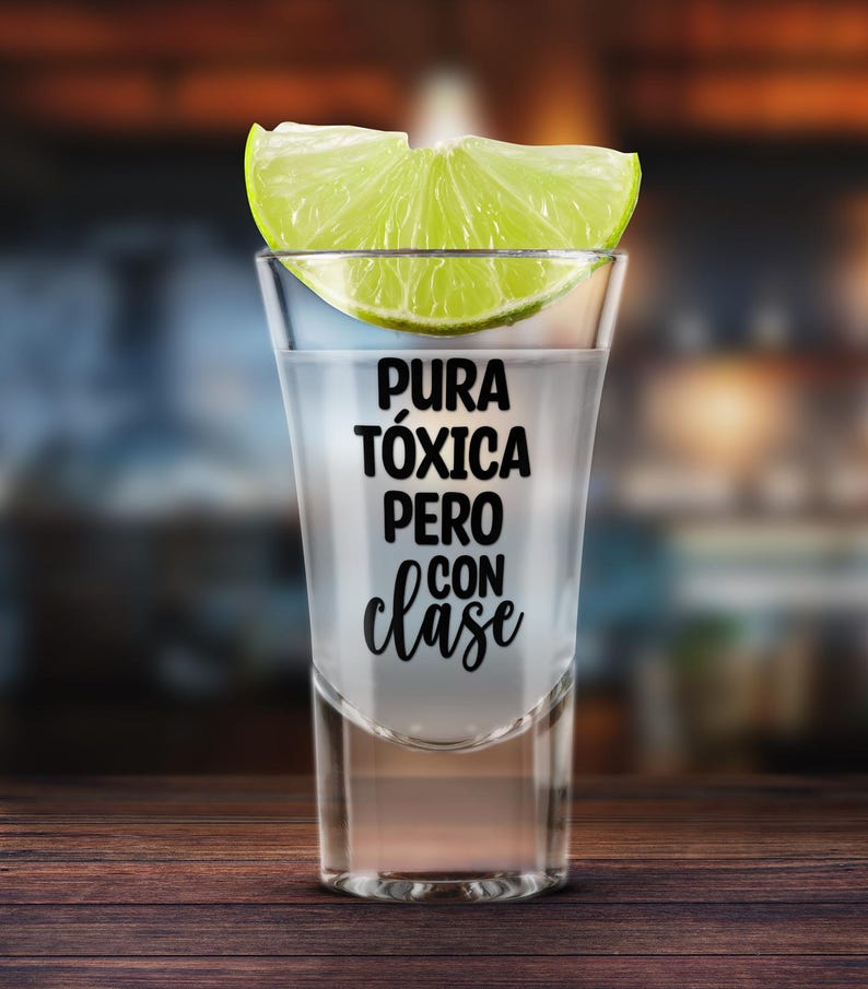 K&ouml;nnte beinhalten: Ein Shotglas gef&uuml;llt mit einer klaren Fl&uuml;ssigkeit, garniert mit einer Limettenscheibe. Auf dem Glas steht der Schriftzug "Pura T&oacute;xica Pero Con Clase". Das Shotglas steht auf einer Holzoberfl&auml;che.
