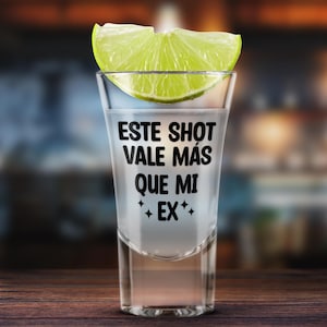 K&ouml;nnte beinhalten: Ein klares Schnapsglas, gef&uuml;llt mit einer klaren Fl&uuml;ssigkeit, garniert mit einer Limettenscheibe. Auf dem Glas steht der Text "ESTE SHOT VALE M&Aacute;S QUE MI EX" in Schwarz. Auf einer Holzoberfl&auml;che.