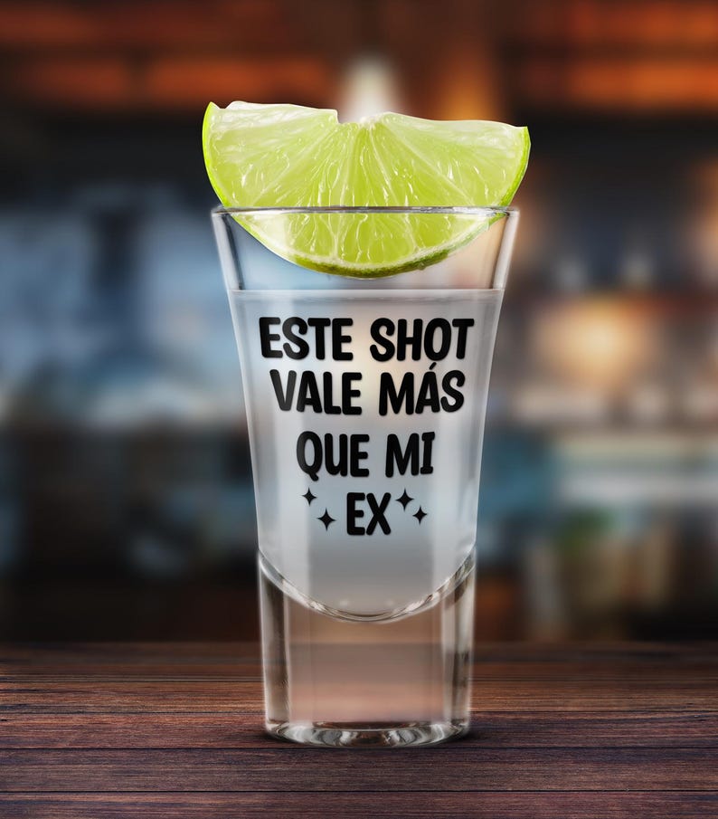 K&ouml;nnte beinhalten: Ein klares Schnapsglas, gef&uuml;llt mit einer klaren Fl&uuml;ssigkeit, garniert mit einer Limettenscheibe. Auf dem Glas steht der Text "ESTE SHOT VALE M&Aacute;S QUE MI EX" in Schwarz. Auf einer Holzoberfl&auml;che.