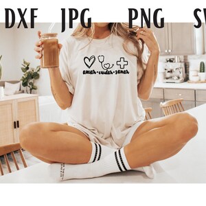 May include: A white t-shirt with a heart, stethoscope, and cross graphic and the text "amar - cuidar - sanar". The model is wearing the shirt and holding a glass of iced coffee.