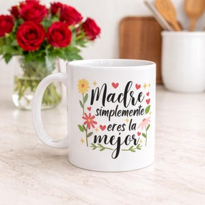 May include: White ceramic mug with the text "Madre simplemente eres la mejor" in black script, decorated with floral and heart accents. A bouquet of red roses is in the background.