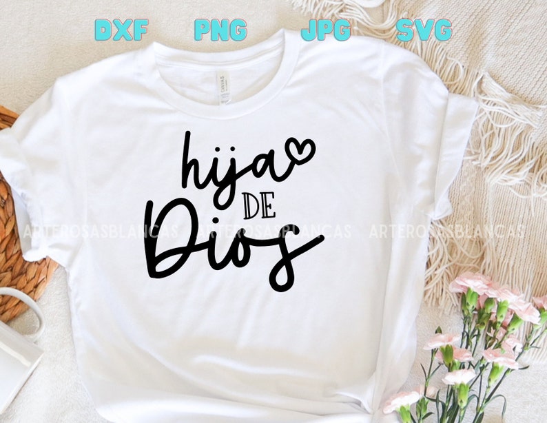 Puede incluir: Camiseta blanca con texto negro que dice "hija de Dios" con un peque&ntilde;o coraz&oacute;n rosa sobre la palabra "hija".